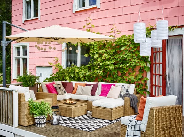 Canapé d'angle, fauteuil et repose-pieds SOLLERÖN en brun et coussins KUNGSÖ blancs sur une terrasse extérieure sous un gran parasol devant une maison rose.