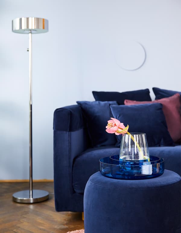 Canapé en velours bleu avec pouf assorti placé devant et lampadaire rond métallique placé à côté, plus des vases en verre colorés de forme arrondie posés sur une table basse, le tout de chez IKEA.