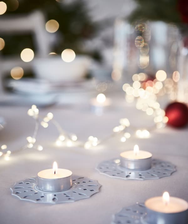 Candeas de té sobre soportes decorativos colocados nunha mesa; en segundo plano, desenfocada, unha gran mesa para a cea de Nadal.