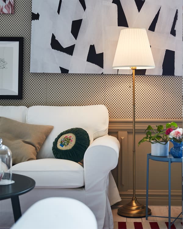 Candeeiro de pé branco, em latão, aceso e perto de um sofá EKTORP branco numa sala.