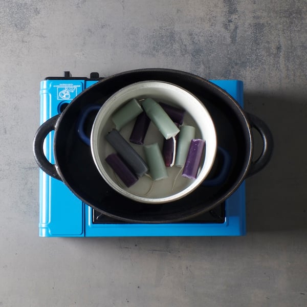 Candle remnants in various colors are heated in a small pot au-bain-marie inside a larger pan, as if they were being melted down for an IKEA-style DIY candle project.