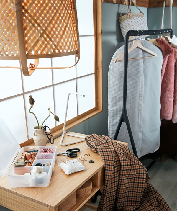 Canto de um quarto organizado para remendar roupa: conjunto de costura e um casaco numa mesa de apoio ao lado de um suporte para cabides com roupa que aguarda a sua vez.