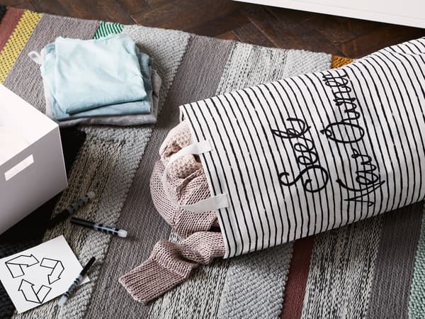 Canvas tote bag embroidered with the words ‘Seek new owner’ and clothes inside lying on a rug next to a box, pens and recycling label.