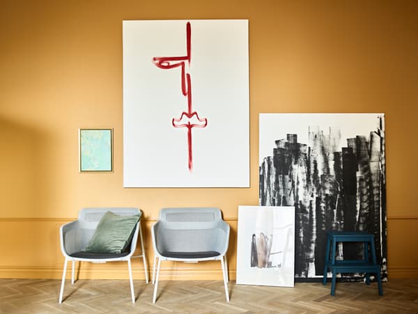 Canvases hanging and leaning on an ochre wall with black and red brushstrokes on them and two chairs in front of them.