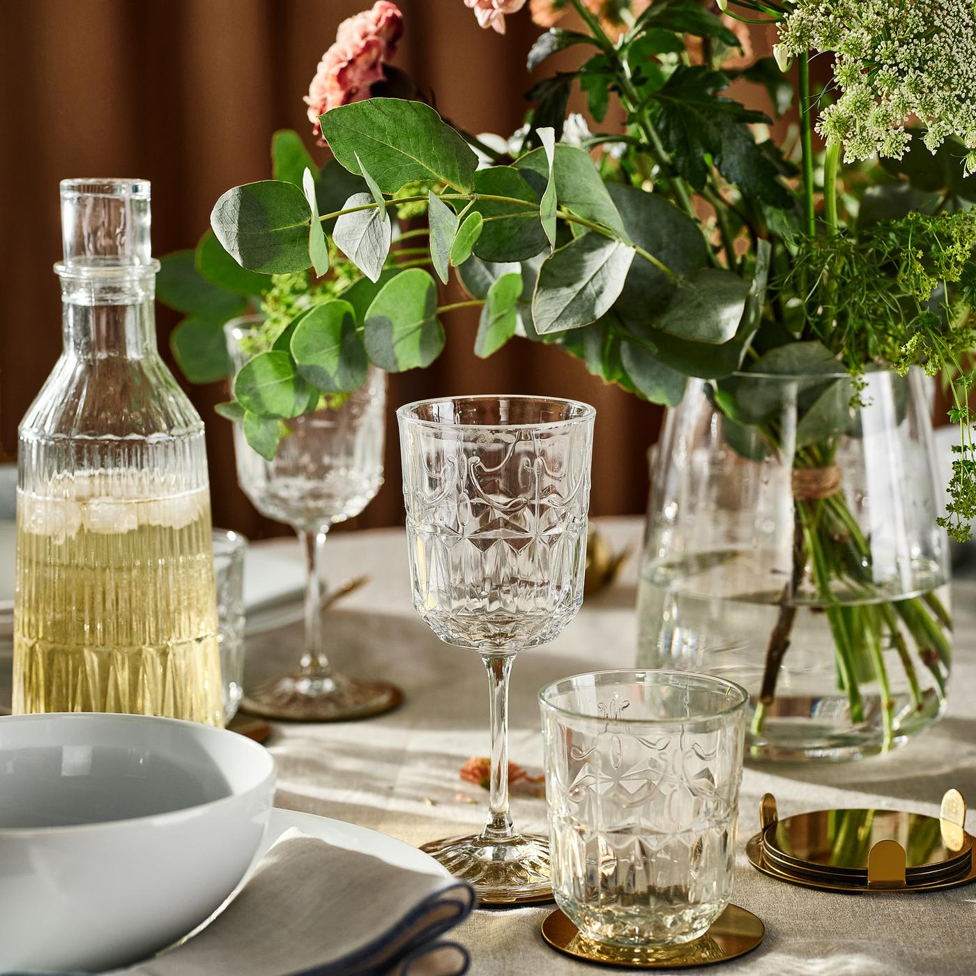 Carafe en verre SÄLLSKAPLIG, verres, verres à vin et vase en verre avec fleurs sur une table ornée d’une nappe de couleur neutre.