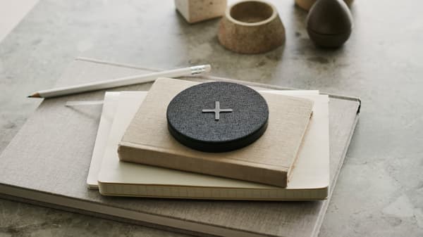 Cargador sen fíos NORDMÄRKE co símbolo "máis" en grande na súa funda téxtil gris sobre unha morea de libros que están enriba dunha mesa.