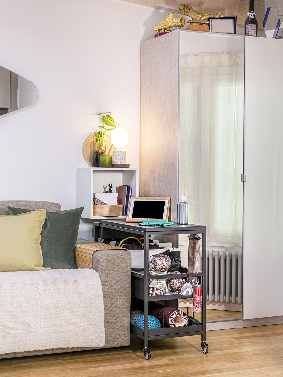 Carrello in antracite TROTTEN contenente diversi materiali in un soggiorno con un divano e un mobile bianco con ante diverse di vetro e di legno. Appeso alla parete un mobile EKET. 