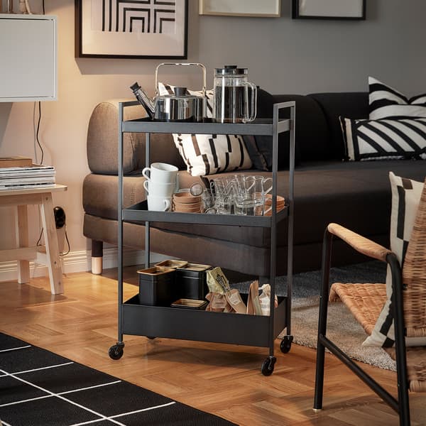 Carrello NISSAFORS nero con un bollitore, delle tazze e delle bustine di tè, posizionato accanto a un divano e a una poltrona - IKEA