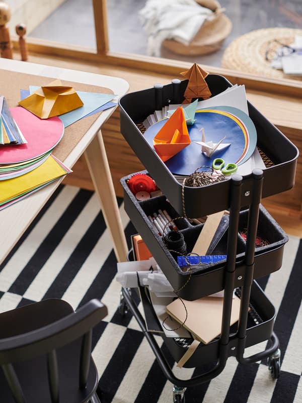 Carrinho RÅSKOG, em preto, com acessórios para trabalhos manuais, junto a um tampo de mesa coberto com papel colorido espalhado.