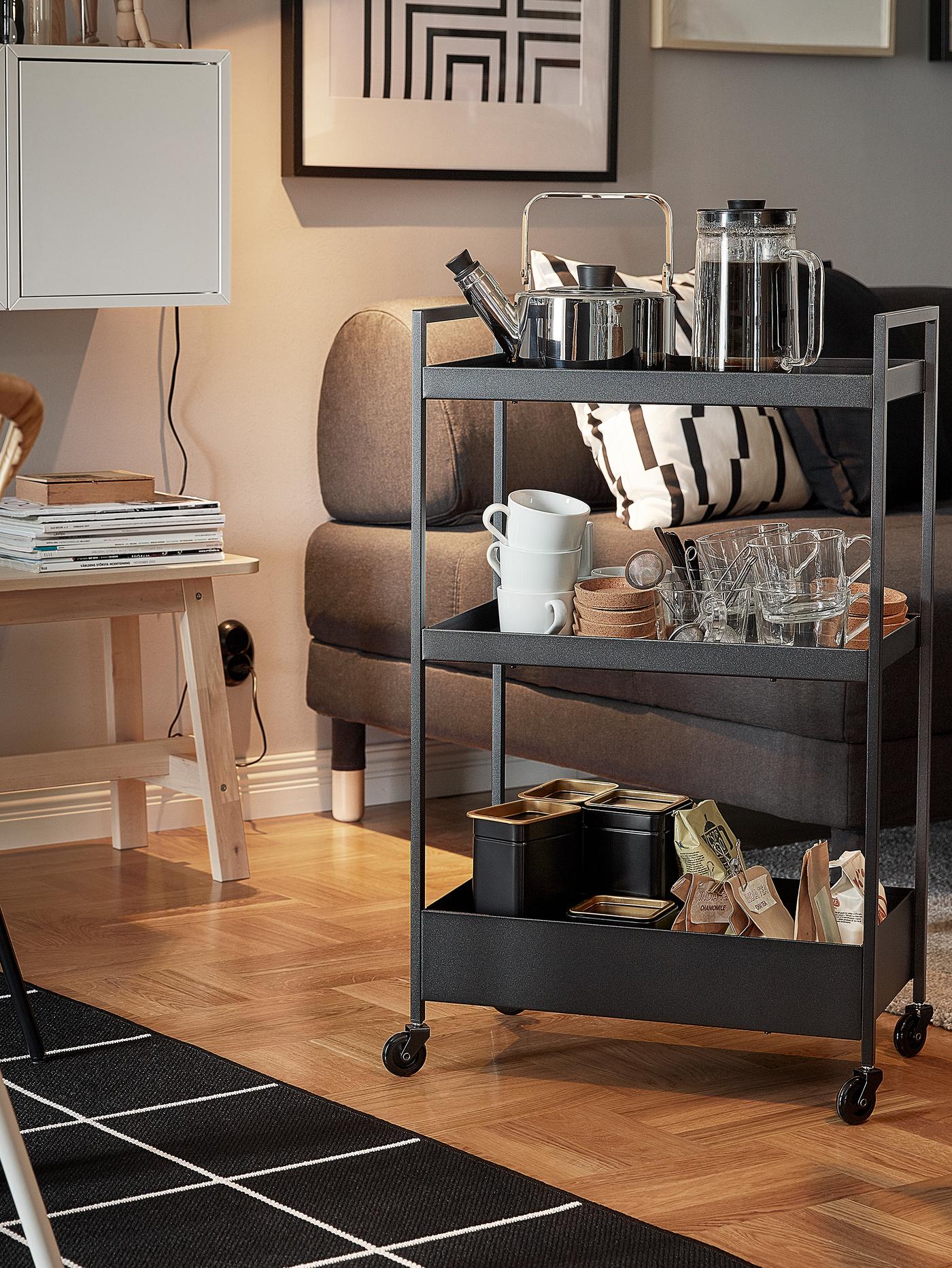 Carrito NISSAFORS negro, situado junto a un sofá y un sillón, con tetera, tazas y bolsitas de té en los estantes.