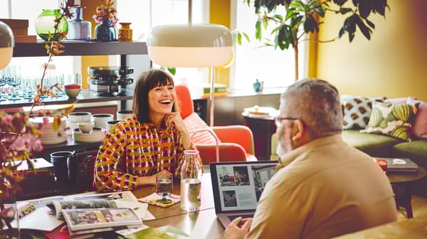 Casal sentado frente a frente numa mesa MÖRBYLÅNGA. Atrás, estante IDÅSEN em metal com copos.