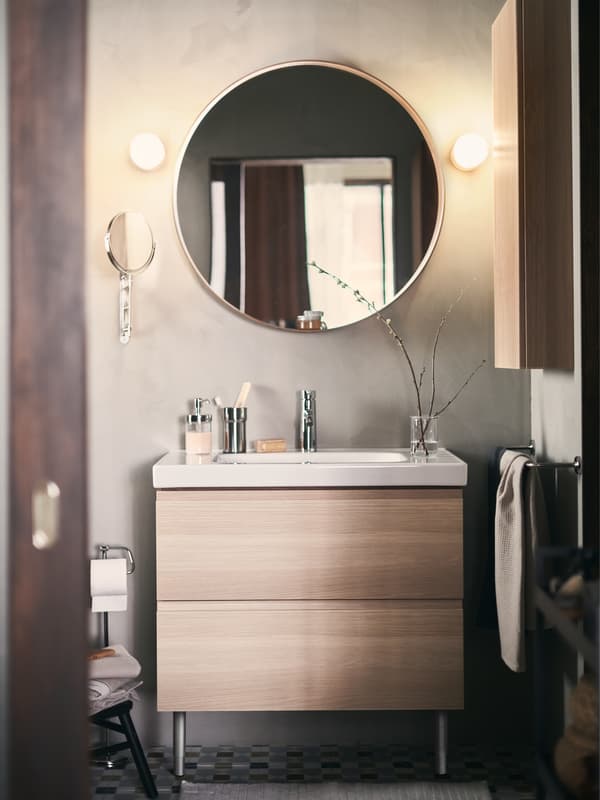 Cermin bulat tergantung di atas kabinet bawah sink dengan dua laci GODMORGON/ODENSVIK. Dua lampu dinding terletak di kiri kanan cermin itu.