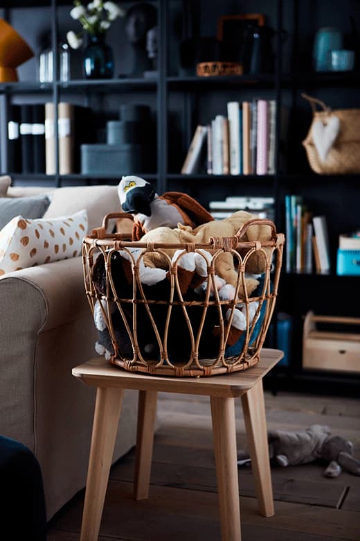 Cesto de verga em cima de um banco na sala, repleto de diferentes peluches de animais.