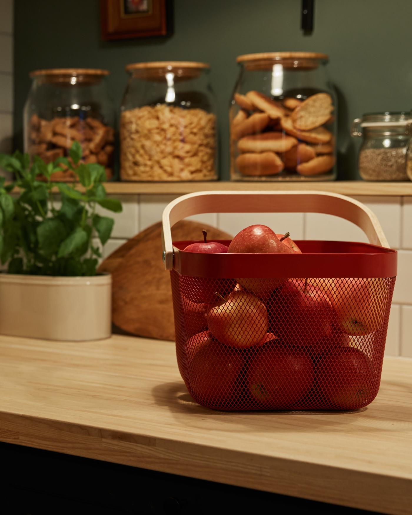 Cesto vermelho numa bancada de cozinha repleto de maçãs vermelhas. Em segundo plano, vários frascos de vidro com comida.