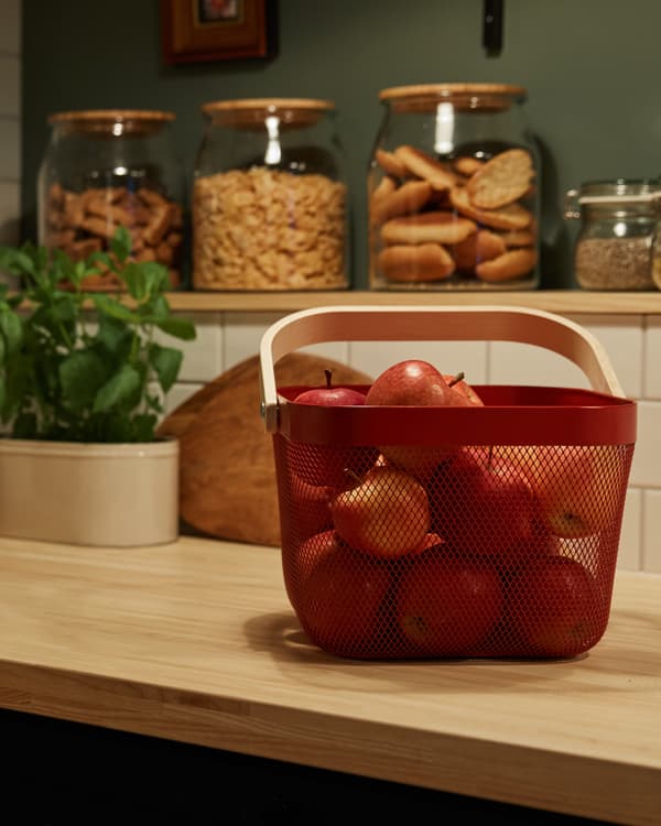 Cesto vermelho numa bancada de cozinha repleto de maçãs vermelhas. Em segundo plano, vários frascos de vidro com comida.