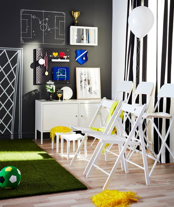 Chairs and stools arranged like stands next to a patch of indoor grass, as prepared for supporters watching football.