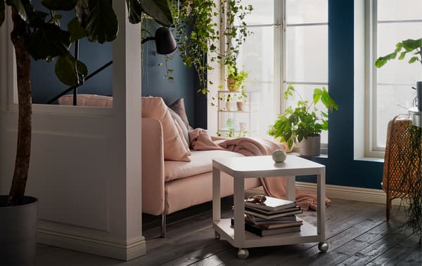 Chaise longue ao sol, janelas altas. Almofadas, manta e candeeiro de leitura. Plantas no parapeito, penduradas e em prateleiras na parede.