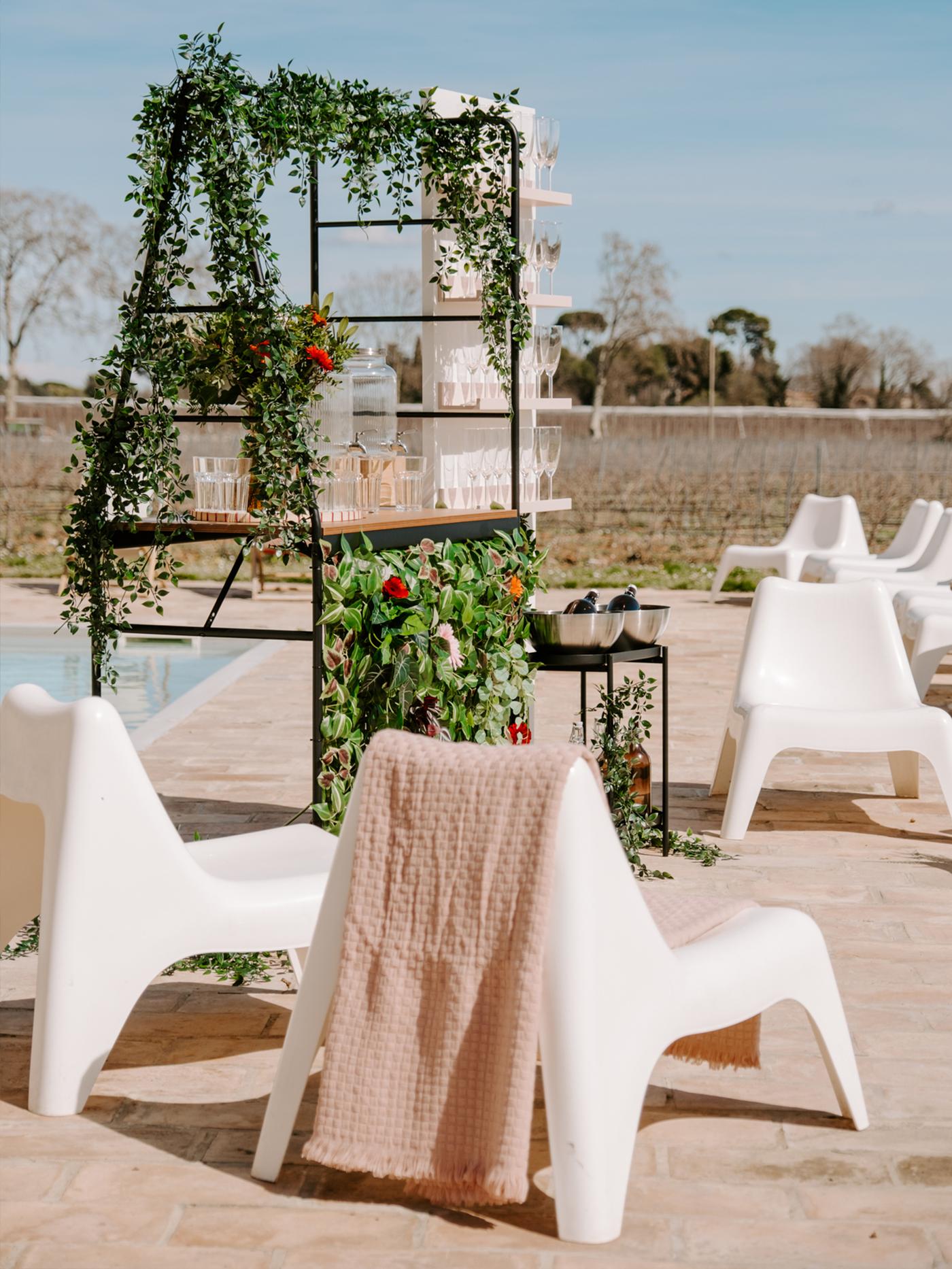 Chaises d'extérieur blanches en plastique à côté d'une piscine avec un bar décoré de fleurs suspendues et de verres.