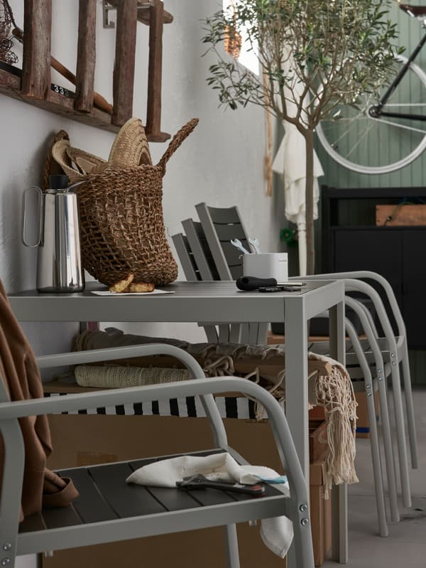 Chaises d’extérieur, une solitaire et d’autres empilées contre un mur, avec une table d’extérieur sur laquelle sont posés une cafetière, un sac et des outils.