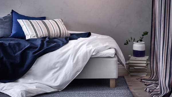 Chambre à coucher avec lit défait, housse de couette ÄNGSLILJA, couvre-lit INDIRA en bleu foncé et 3 coussins.