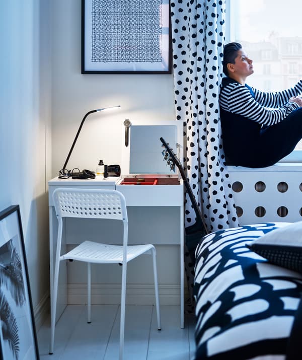 Chambre à coucher avec rideaux à pois noirs, bureau, chaise et plancher blancs. Une femme est assise sur l'appui de fenêtre.