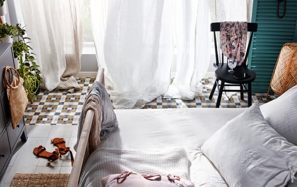 Chambre à coucher avec une grande fenêtre ouverte devant laquelle sont suspendus des voilages qui ondulent sous la brise.