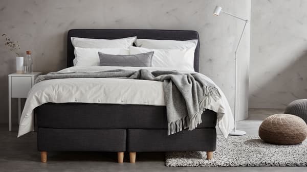 Chambre à coucher gris clair avec lit/sommier DUNVIK gris foncé, linge de lit blanc, lampadaire liseuse NYMÅNE en blanc, et deux poufs.
