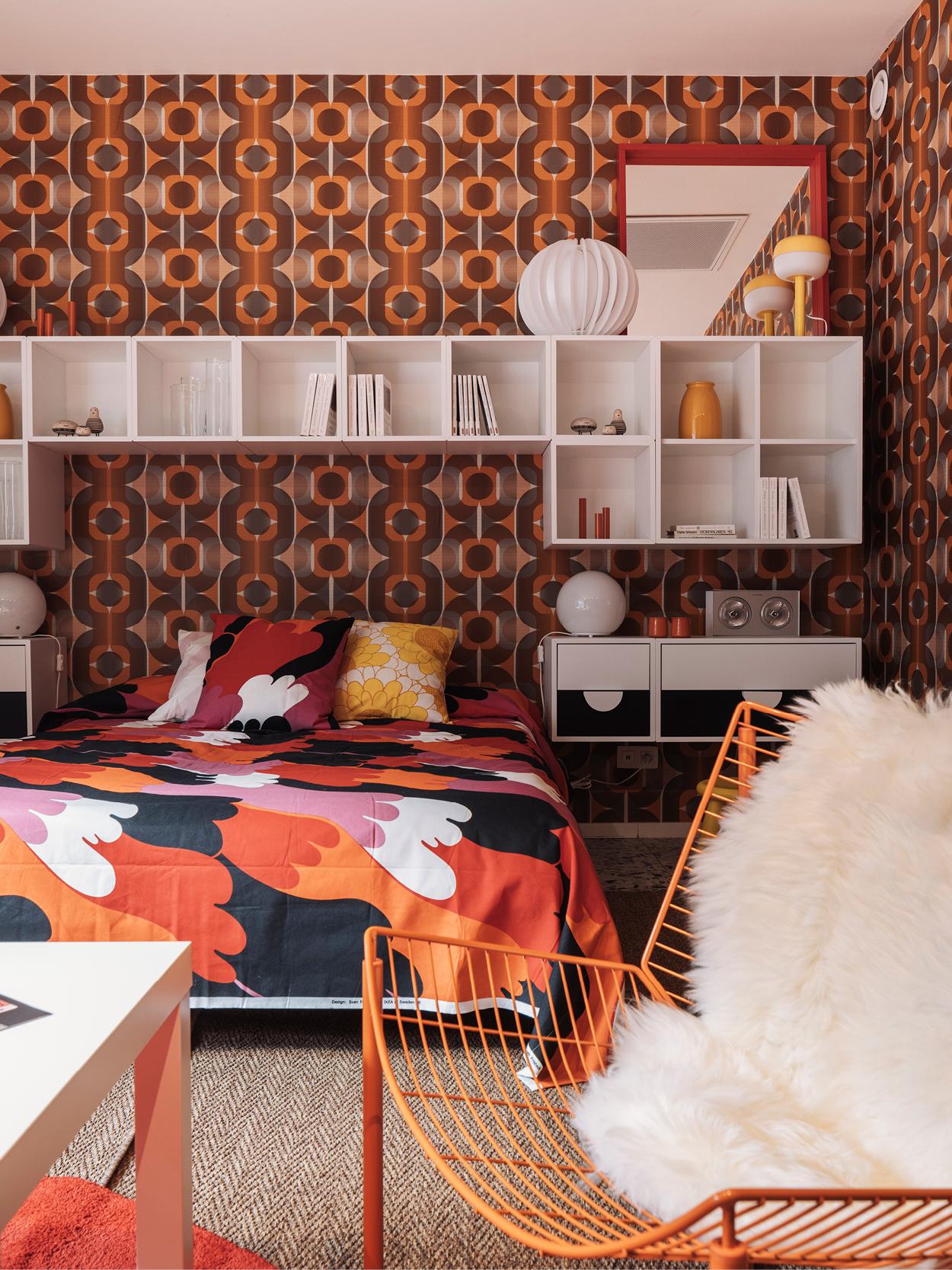 Chambre au style rétro avec une literie colorée, du papier peint géométrique orange et marron, des étagères blanches et une chaise en fil orange avec un coussin en peau de mouton blanche.