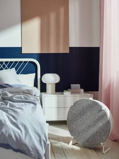 Chambre confortable avec cadre de lit en métal blanc, linge de lit bleu clair, table de chevet blanche avec lampe et livres, murs bleu marine et blancs, rideaux roses et enceinte ronde grise.