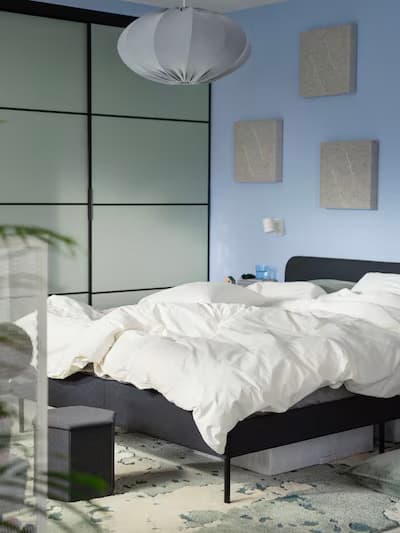 Chambre moderne avec linge de lit blanc, tête de lit noire, mur bleu clair, garde-robe coulissante en verre dépoli et suspension blanche.