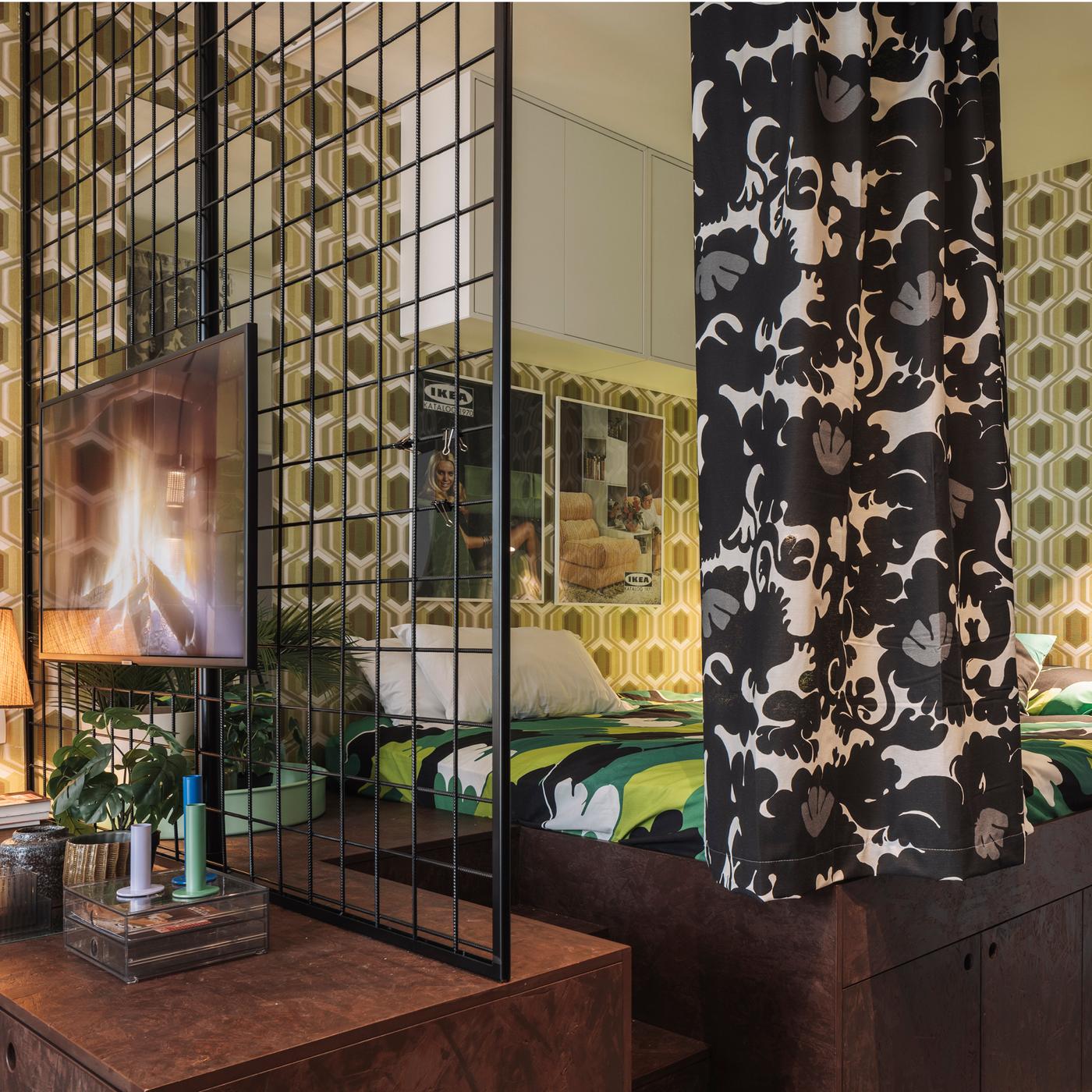 Chambre rétro avec literie verte et blanche, rideau floral noir et blanc, papier peint géométrique dans les tons de vert, grille métallique avec télévision montée et une armoire en bois avec des bougies et des plantes.