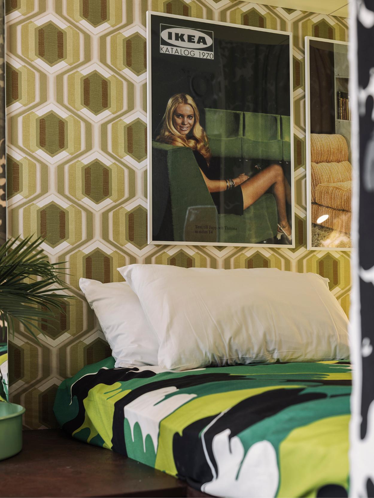 Chambre rétro avec papier peint géométrique dans les tons de vert et de marron, une affiche du catalogue IKEA de 1970 sur le mur, un lit avec une literie blanche et verte et une plante dans un pot vert à côté du lit.