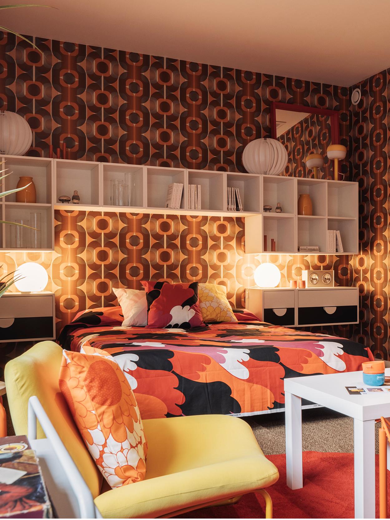 Chambre rétro avec papier peint géométrique en marron, orange et jaune, literie colorée, tables de chevet avec lampes rondes, étagère murale avec décoration et une chaise jaune avec coussin orange sur un tapis rouge.