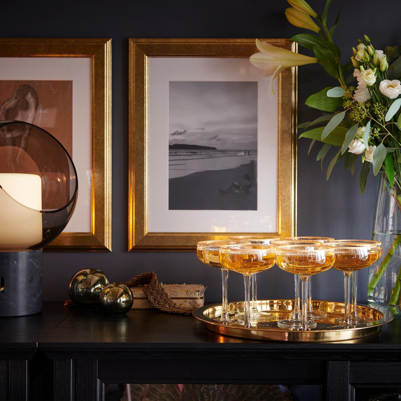 Champagne glasses on a tray, in front of a flower arrangement in a glass vase and two gold-framed pictures on the far wall.