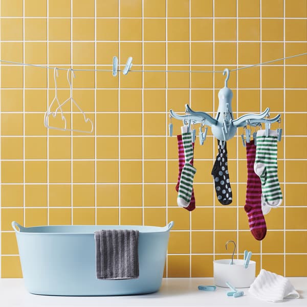 Chaussettes sur un séchoir à suspendre bleu, pinces et cintres accrochés à un fil à linge au-dessus d’un panier à linge bleu, sur fond de mur carrelé jaune.
