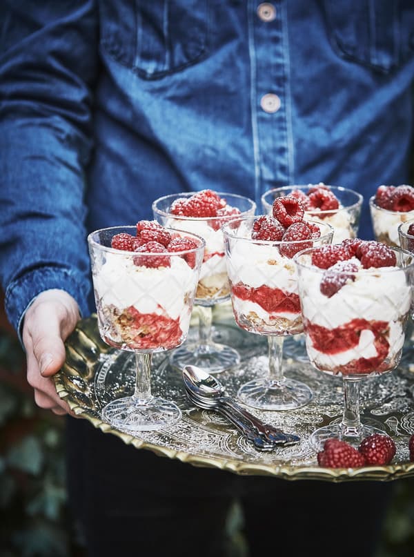 Cheesecake and raspberries are layered in glasses and carried on a silver tray.