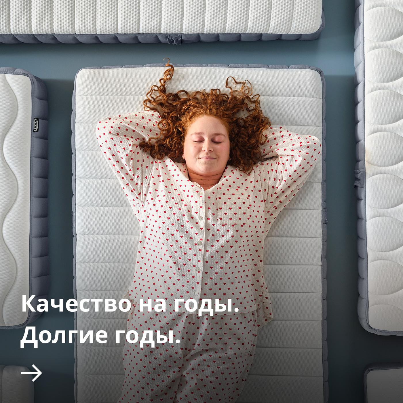 Человек в пижаме с узором лежит на матрасе, руки за головой, вид сверху, в окружении других матрасов.
