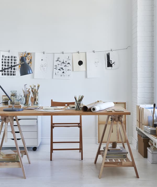 Chez un artiste, plateau de bureau en bois posé sur deux tréteaux et dessins suspendus à un câble placé au mur.