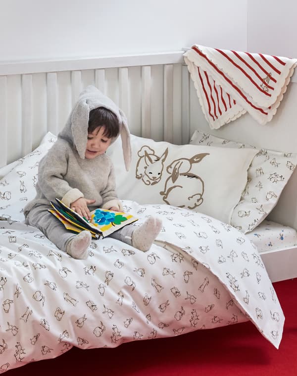 Child in a onesie with rabbit ears sitting in a crib with RÖDHAKE rabbit themed bed linen reading a child’s picture book.