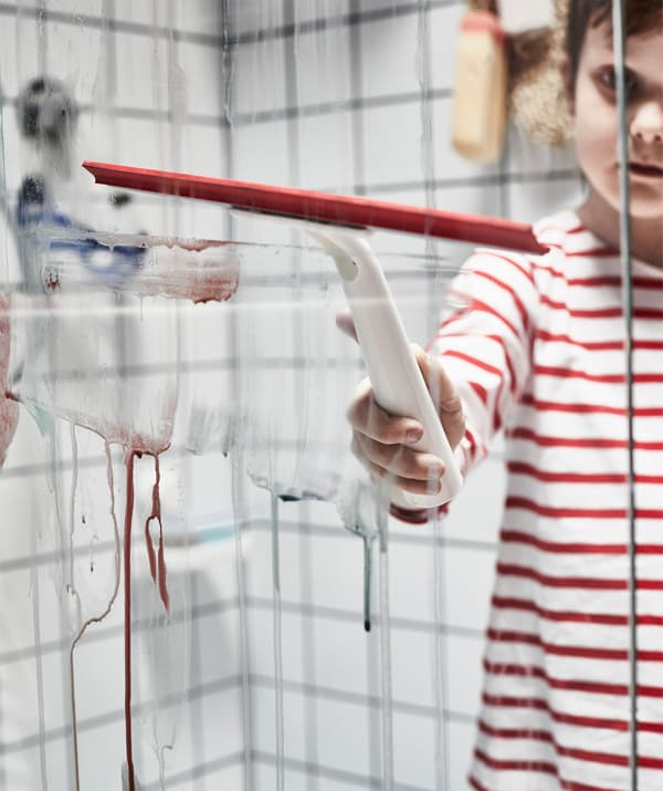 Child in a striped shirt standing inside a dry shower, using a squeegee to clean a glass partition from watercolours.