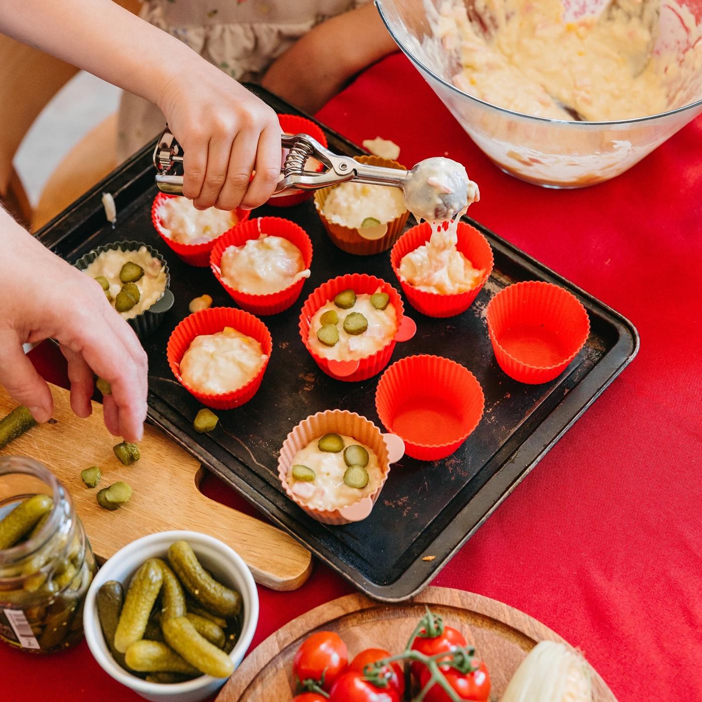 Child scooping batter into silicone muffin cups on a baking tray, topping with pickle slices, with a bowl of mixture and fresh vegetables nearby.
