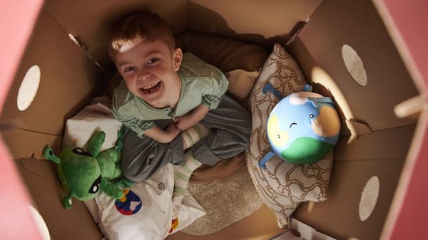 Child sitting inside a cardboard playhouse with an Earth pillow and green alien plush toy.