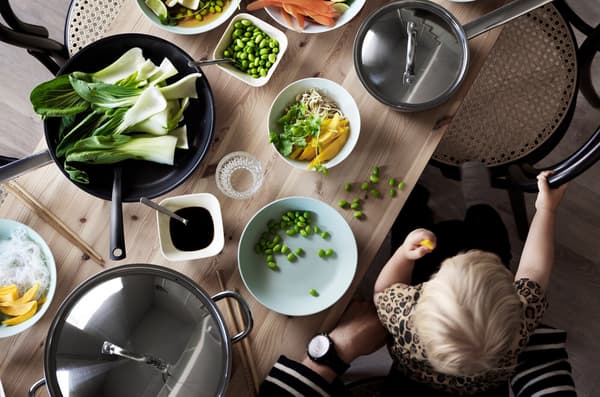 Child sitting on adult’s lap eating finger food at a wooden table with sliced fruit and vegetables in bowls and saucepans.