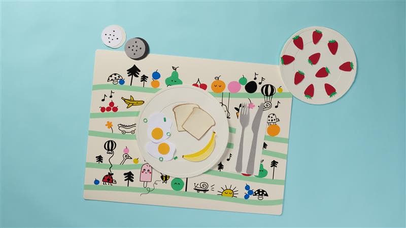 Children's placemat with plate on top containing 2 eggs, a banana, and 2 pieces of toast. A plate with a strawberry pattern sits on the top right of the placemat.