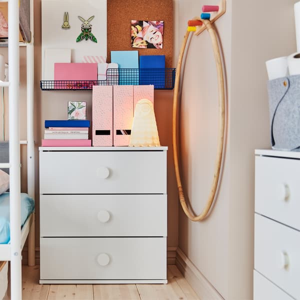 Children's room with two white chests of drawers that have books, a white wall lamp and pink magazine files standing on top.