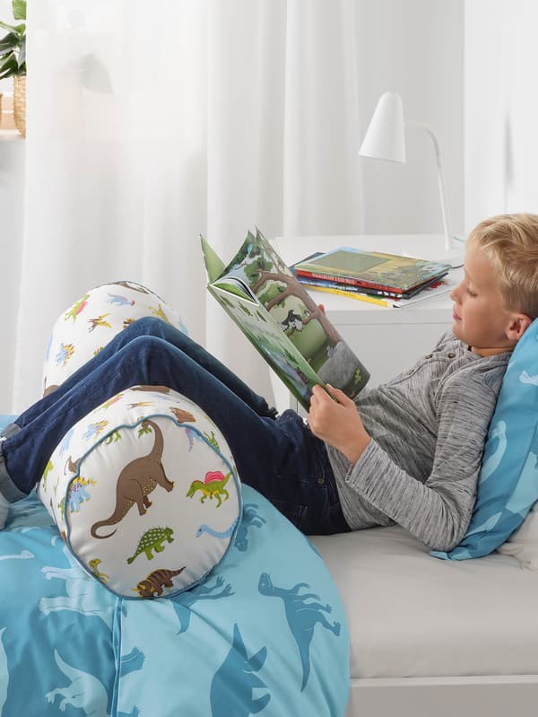 Chlapec si číta v posteli s modrou posteľnou bielizňou JÄTTELIK, nohy má opreté o vankúš JÄTTELIK, v pozadí je biely záves a biela lampa FUBBLA na bielej komode MALM.