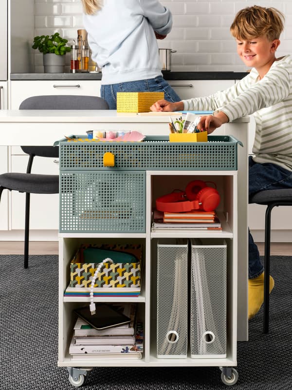 Chlapec si kreslí pri bielom jedálenskom stole, vedľa stola je vozík ÖVNING, v pozadí je mamička, ktorá varí.