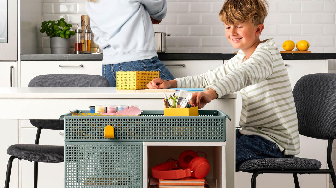 Chlapec si kreslí pri bielom jedálenskom stole, vedľa stola má vozík ÖVNING, v pozadí je mamička, ktorá varí.