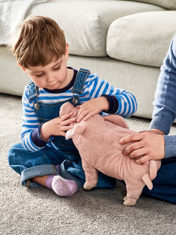Chlapec v pruhovanom tričku sedí na sivom koberci pred sivou pohovkou a hrá sa s plyšovým prasiatkom KNORRIG.