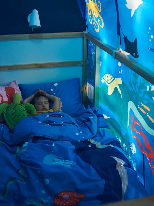 Chlapec zaspáva v posteli v modrej posteľnej bielizni BLÅVINGAD a baldachýnom KURA, vedľa neho leží plyšová korytnačka BLÅVINGAD.